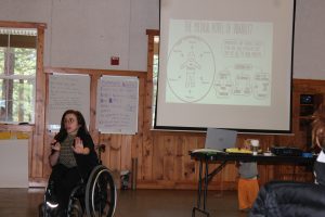 Youth in a wheelchair speaking into a microphone with a slideshow behind her.