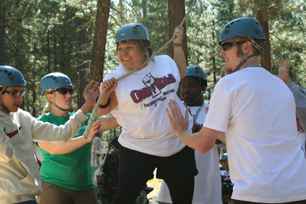 Photo of Dolores uses a rope to balance during the ropes course. Her team stands around her for support.