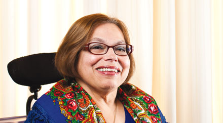Photo of a white woman with shoulder-length auburn hair  in a wheelchair with colorful scarf and eyeglasses smiling.